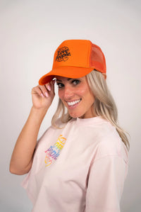 Woman wearing an orange cap with text and a light pink hoodie with colorful graphics on a plain background