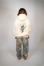Person wearing a beige hoodie with a hand gesture design and patterned pants on a plain background